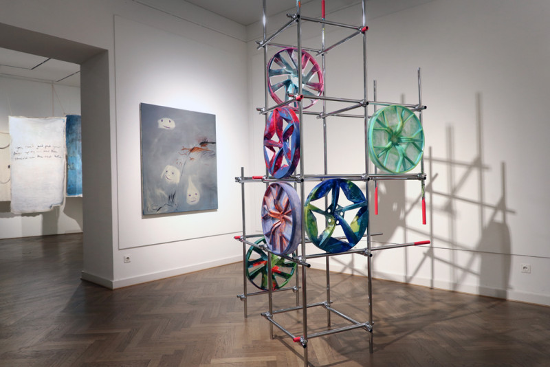 Eine Ansicht des Ausstellungsraum. Im Vordergrund ist eine Installation aus Mteallstangen und bunten Autofelgen zu sehen, im Hintergrund ein grau blaues Bild sowie ein offener Durchgang zu einem weiteren Raum.