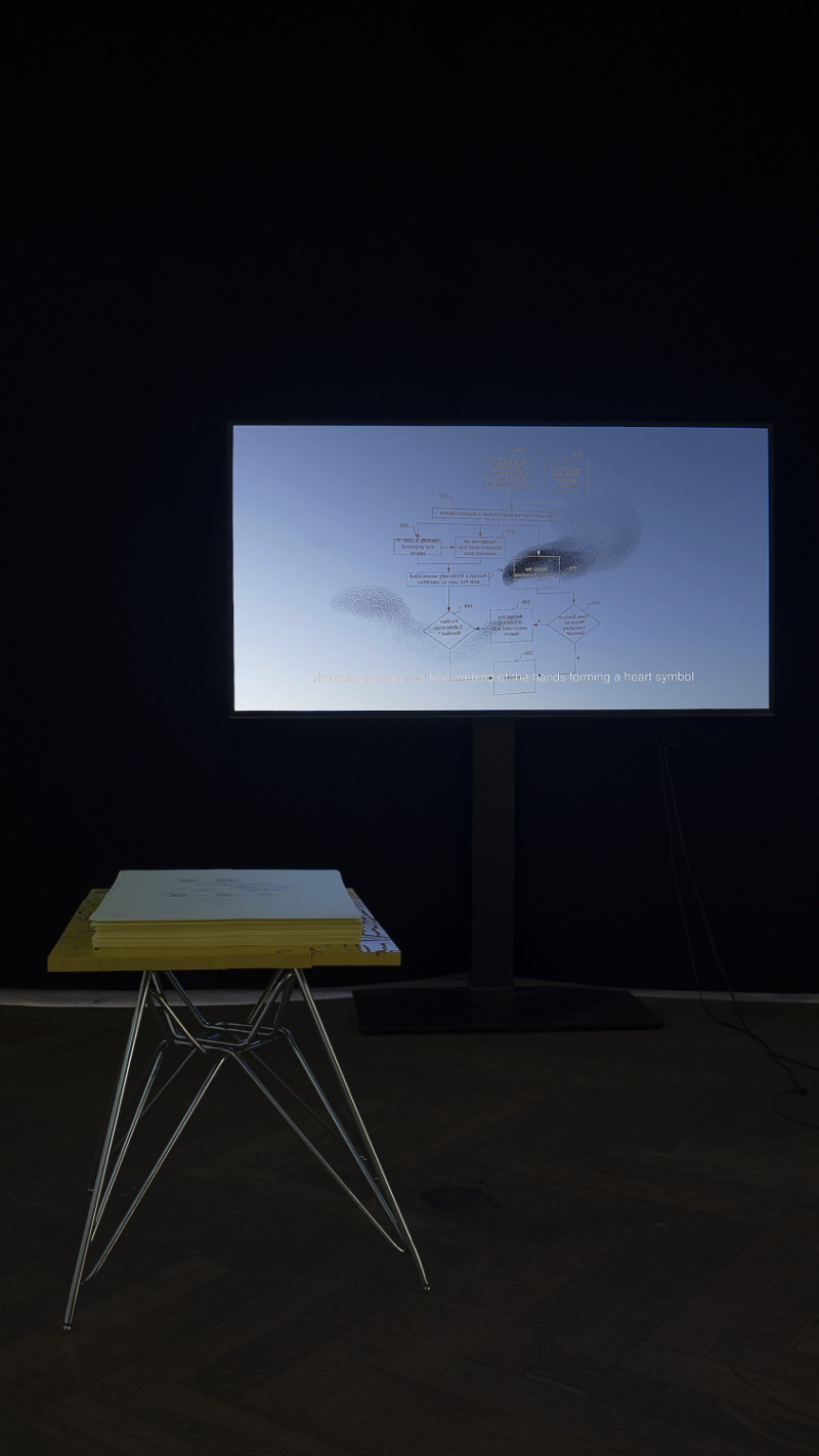 Dunkle Raumansicht. Dort steht ein metallener Tisch mit gelber Tischplatte und einem Papierstapel darauf. Ein blau-leuchtender Bildschirm zeigt eine Erklärungsgrafik..