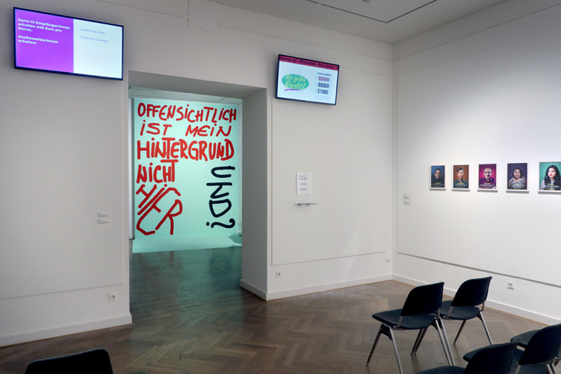 Blick durch die Räumlichkeiten. Es sind hochangebrachte leuchtende Bildschirme und Stühle zu sehen, Portraitbilder und das blaue Plakat mit rot-schwarzer Schrift.