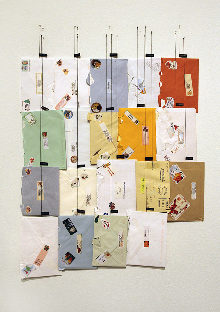 19 Briefe in verschiedenfarbigen Umschlägen hängen in einem Block an Schnüren befestigt an der Wand.