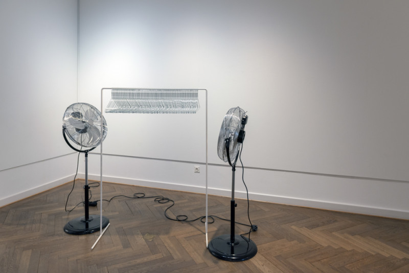 Zwei Ventilatoren stehen sich gegenüber. Dazwischen steht eine Art Kleiderständer, auf dem 98 Metallkleiderbügel hängen.