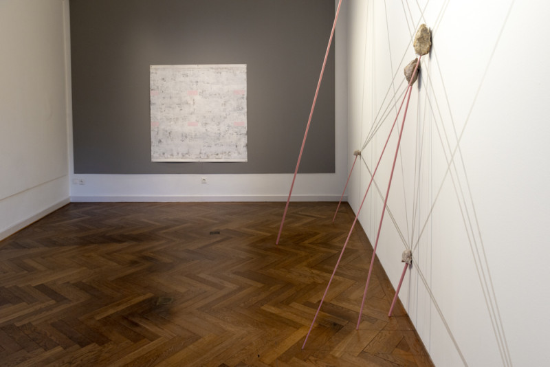 Ein abstraktes in Rosa- und Grautönen gehaltenes Gemälde hängt an einer grauen Wand. Dünne rosafarbene Metallstangen sind an die Wand gelehnt und scheinen Steine darauf zu stützen.