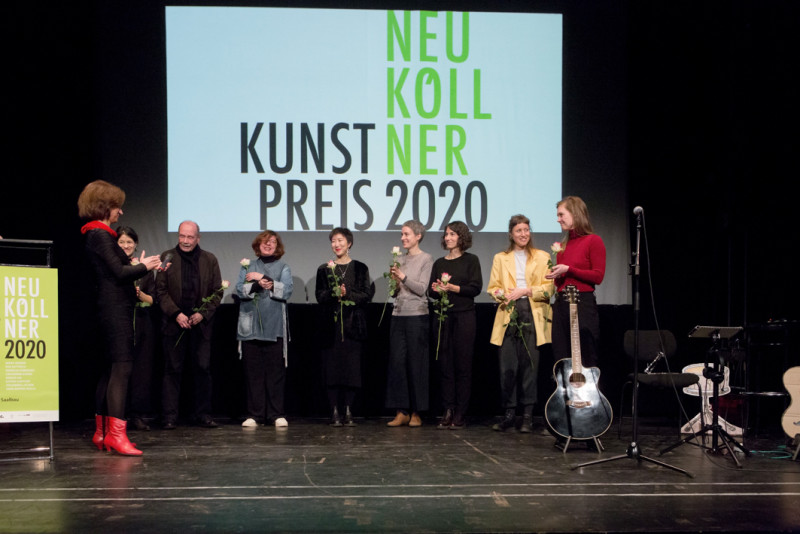 Die acht nominierten Künstler:innen stehen auf der Bühne vor der Projektion des Neuköllner Kunstpreis-Logos mit jeweils einer Rose in der Hand.