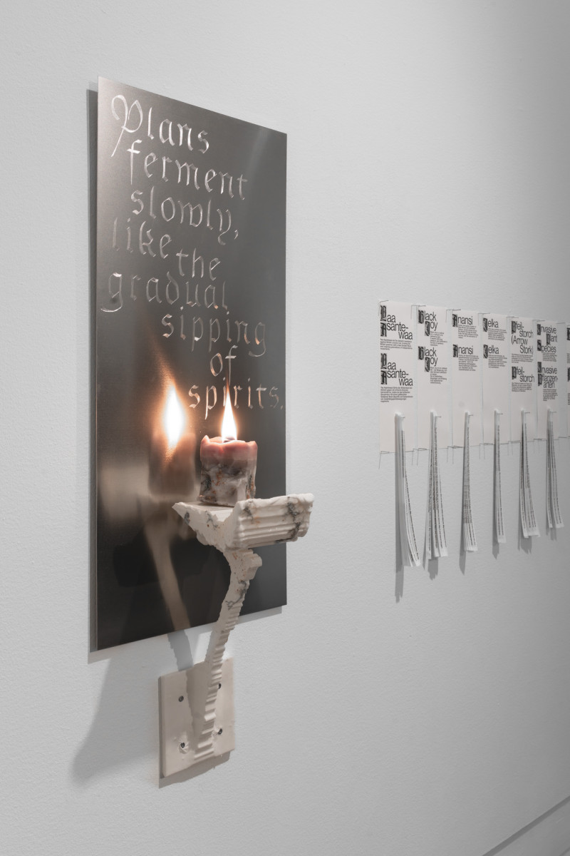 Man sieht eine silberne Platte an der Wand hängen mit einem eingravierten Spruch. Davor ist mit einer weißen Konstruktion eine brennende Kerze angebracht. Im Hintergrund hängen Papierfetzen von der Wand.