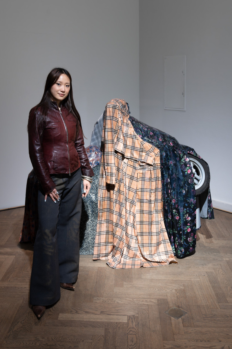 Man sieht die Künstlerin vor ihrer Skulptur stehen. Die Skulptur besteht aus einem Motorrad über das Textilien gelegt sind. Von dem Motorrad ist lediglich das Vorderrad zu erkennen.