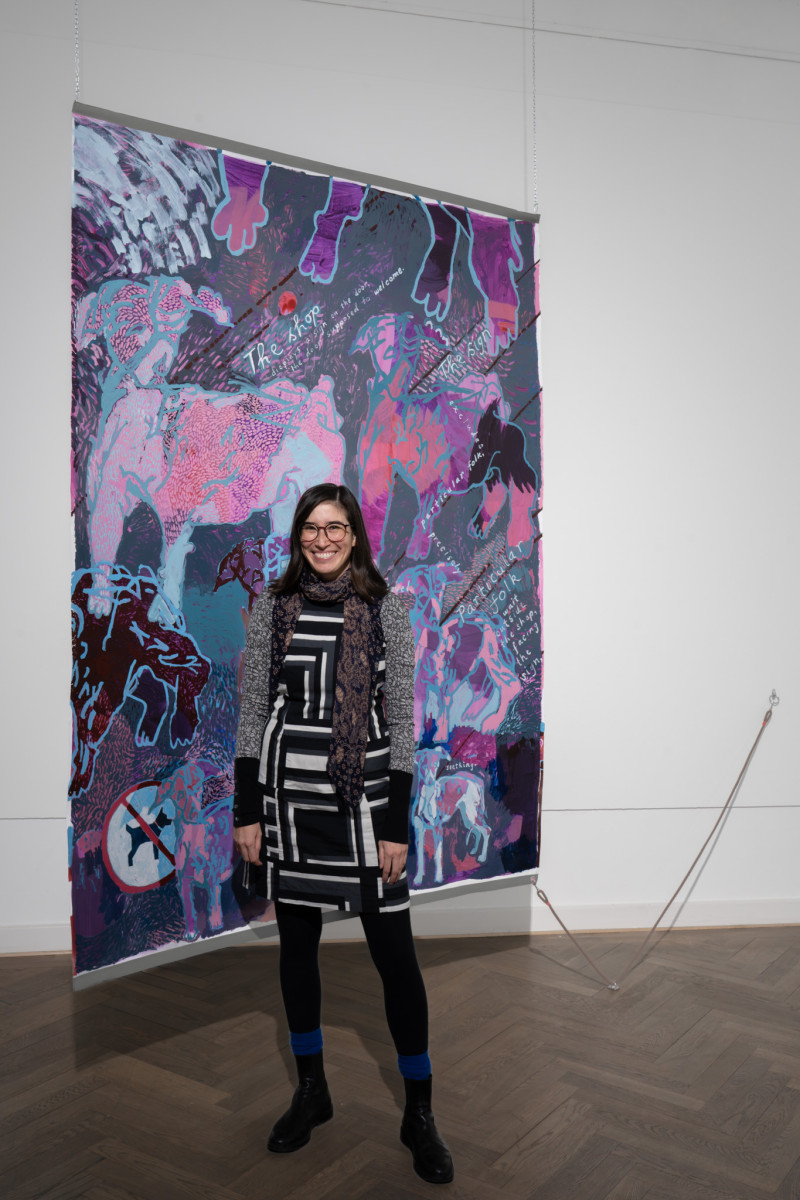 Die Künstlerin steht vor ihrem rosa-lila Gemälde mit einem Hund drauf.