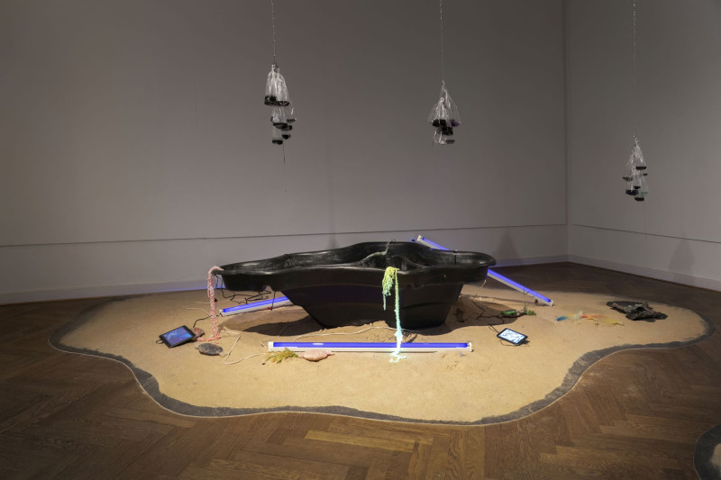 Zwei Neonröhren mit Schwarzlicht liegen auf einem Sandhaufen neben einem Gartenteich-Schale und mehreren Ipads mit Videoarbeiten.